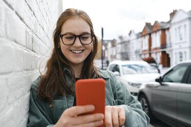 Lächelnde Frau mit Smartphone an die Wand gelehnt - ASGF03547