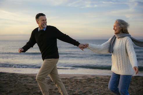 Cheerful mature couple holding hands and running at beach - EBSF03141
