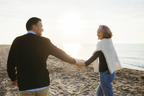 Lächelnde reife Frau hält Hände mit Mann und geht am Strand spazieren - EBSF03177