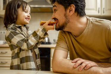 Happy boy feeding pizza to father at home - ANAF01198