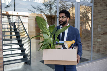 Thoughtful businessman carrying cardboard box leaving office building - VRAF00086