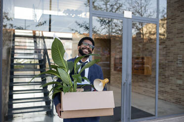 Happy businessman with carrying box with office supplies in front of glass wall - VRAF00087
