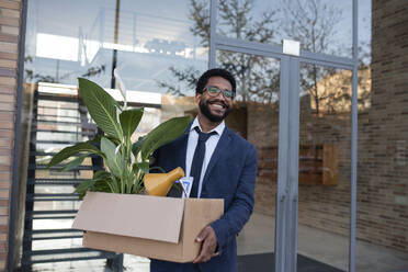 Smiling businessman leaving office building carrying cardboard box - VRAF00088