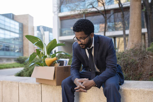 Gestresster Geschäftsmann mit Pappkarton an der Wand sitzend in der Nähe eines Bürogebäudes - VRAF00089