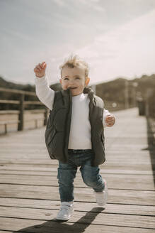 Happy cute son wearing padded jacket running at jetty on sunny day - MTBF01266