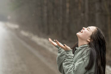 Cheerful teenage girl enjoying rain on road - ANAF01204