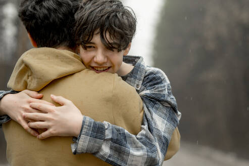 Lächelnder Sohn im Teenageralter umarmt Vater an einem regnerischen Tag - ANAF01209