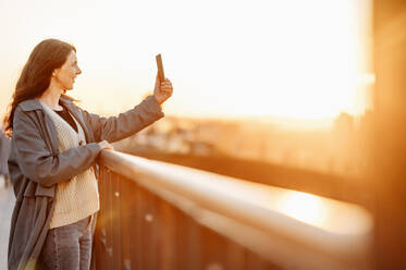 Eine fröhliche Frau fängt den Moment mit einem Selfie auf einer Brücke bei einem wunderschönen Sonnenuntergang ein - ANAF01216