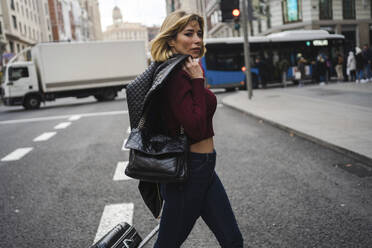 Frau mit Gepäck und Lederjacke beim Überqueren der Straße in der Stadt - JJF00759
