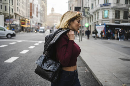 Blonde Frau mit Lederjacke beim Überqueren der Straße in der Stadt - JJF00760