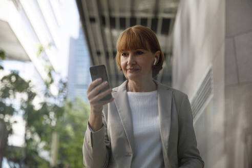 Ältere Geschäftsfrau benutzt Mobiltelefon vor einem Gebäude - IKF00087