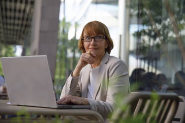 Nachdenkliche Geschäftsfrau mit Hand am Kinn, die mit ihrem Laptop in einem Straßencafé sitzt - IKF00095