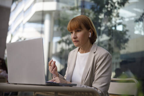 Businesswoman wearing wireless in-ear headphones doing video call through laptop - IKF00100