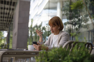 Businesswoman doing video call through smart phone sitting at sidewalk cafe - IKF00103