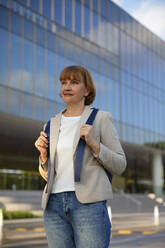 Nachdenkliche Geschäftsfrau mit Rucksack vor einem Gebäude stehend - IKF00110
