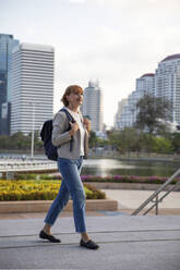 Mature businesswoman wearing backpack walking on footpath - IKF00123