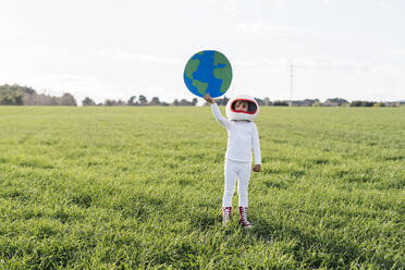 Girl wearing astronaut costume holding cut out of earth standing on grass - JCZF01214