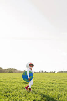 Girl wearing astronaut costume holding earth cut out standing on grass - JCZF01216