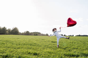 Playful girl holding heart shape balloon standing on grass - JCZF01217