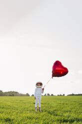 Girl dressed as astronaut holding heart shape balloon standing on grass - JCZF01218