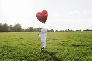 Girl dressed as astronaut walking on meadow holding heart shape balloon - JCZF01219