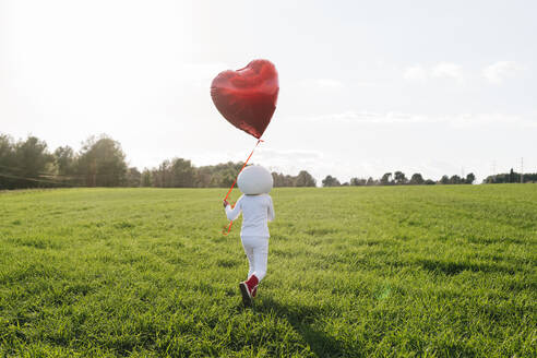 Als Astronautin gekleidetes Mädchen, das auf einer Wiese spazieren geht und einen herzförmigen Luftballon hält - JCZF01219