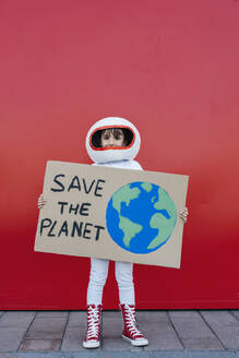 Girl wearing astronaut costume holding Save The Planet placard standing in front of red wall - JCZF01221