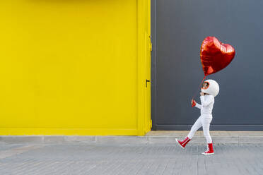 Mädchen hält Herzform Ballon zu Fuß in der Nähe von gelben Wand - JCZF01222