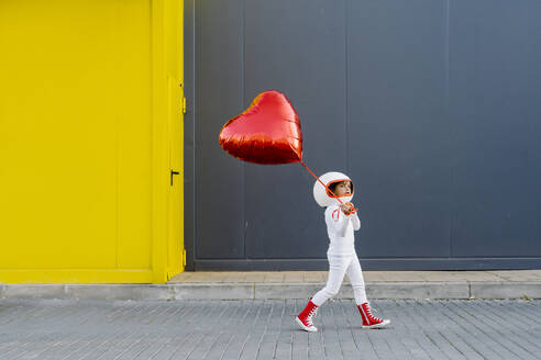 Girl wearing astronaut costume walking with heart shape balloon near yellow wall - JCZF01223