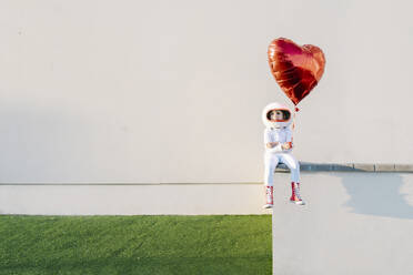 Girl wearing astronaut costume sitting on wall holding heart shape balloon - JCZF01224