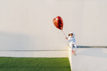 Girl dressed as astronaut sitting on wall holding heart shape balloon - JCZF01225
