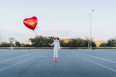 Mädchen im Astronautenkleid steht auf einem Parkplatz und hält einen herzförmigen Ballon - JCZF01231