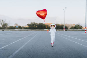 Mädchen im Astronautenkleid geht auf einem Parkplatz und hält einen roten Herzballon - JCZF01236