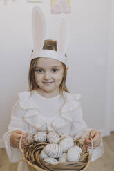 Smiling girl wearing rabbit ears holding basket of Easter eggs at home - VBUF00300