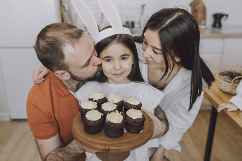 Eltern, die ein Mädchen umarmen, das zu Hause Muffins hält - VBUF00304