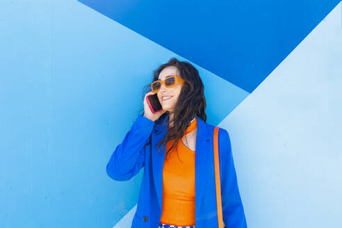 Happy fashionable woman wearing sunglasses talking on smart phone in front of blue wall - EGHF00747