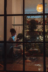 Geschäftsfrau, die nachts auf dem Sofa ein Mobiltelefon benutzt, gesehen durch Glas - JOSEF18452