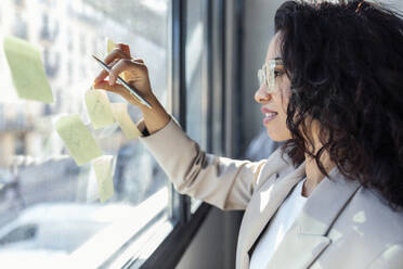 Smiling businesswoman sticking adhesive notes on glass window in studio - JSRF02495