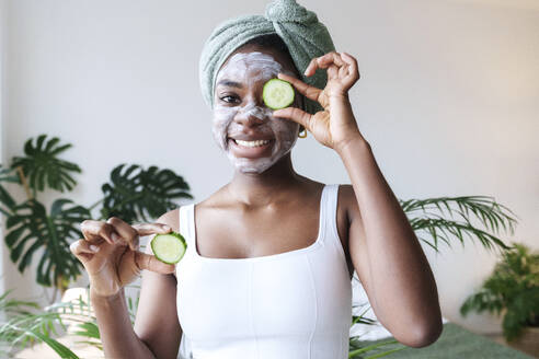 Happy young woman holding cucumber over eye at home - AAZF00291