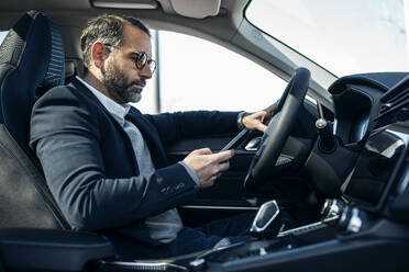 Businessman using smart phone sitting in electric car - JSRF02514