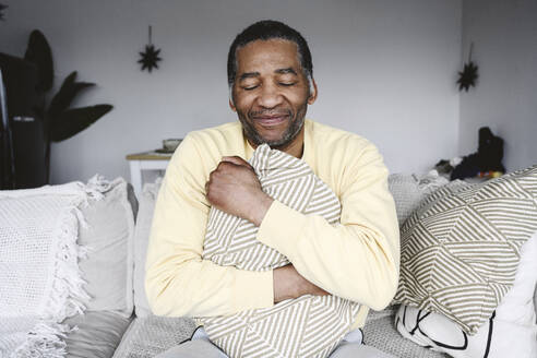 Smiling mature man hugging pillow sitting on sofa at home - EYAF02580