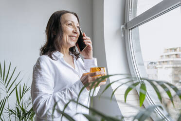 Glückliche Geschäftsfrau, die mit einem Smartphone spricht und ein Glas in der Nähe des Fensters hält - OSF01525