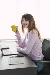 Lächelnde Geschäftsfrau sitzt mit Kaffeetasse am Schreibtisch - PNAF05203