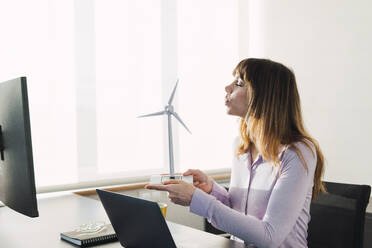 Businesswoman blowing on wind turbine model in office - PNAF05205