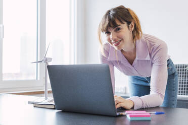 Lächelnde Geschäftsfrau lehnt sich mit ihrem Laptop im Büro an den Schreibtisch - PNAF05223