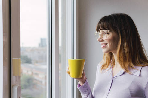 Lächelnde Geschäftsfrau mit Kaffeetasse schaut durch ein Fenster - PNAF05231