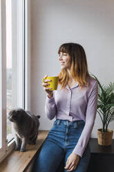 Lächelnde Geschäftsfrau mit Kaffeetasse, die am Fenster sitzt - PNAF05233