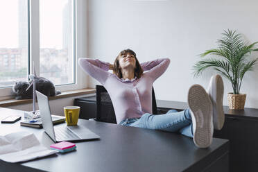 Smiling businesswoman with hands behind back sitting at desk - PNAF05236