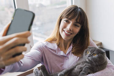 Freudige Frau fängt einen schnurrigen Moment mit ihrem Katzenfreund ein, indem sie ihr Smartphone für ein Selfie benutzt - PNAF05239