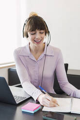 Happy businesswoman wearing headset writing on note pad at desk - PNAF05259